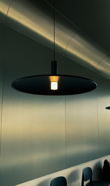 A minimalist, modern interior setting featuring a sleek black pendant lamp hanging from the ceiling. The lamp casts a warm glow, illuminating a section of the room. The background consists of smooth, light-colored walls with a slight metallic sheen, and a row of dark chairs partially visible at the bottom of the image.