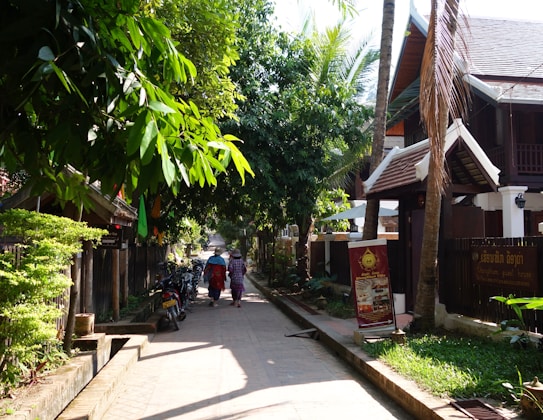 A narrow, tree-lined street with two people walking away, wearing traditional attire and carrying umbrellas. Motorbikes are parked along the side under the shade of lush, green trees. Traditional buildings with wooden accents flank the street, adding to a serene and quaint atmosphere.