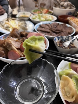 A variety of dishes are set on a table, featuring a close-up view of a dumpling held by chopsticks. Surrounding plates contain meats, seafood, sliced sausage, and fresh salads, creating an abundant and diverse spread.