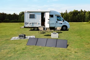 A motorhome is parked on a lush green field surrounded by trees. Nearby, a portable solar panel is set up on the grass beside camping chairs and a table. There is a small power generator and an outdoor rug, creating a comfortable outdoor scene.
