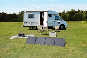 A motorhome is parked on a lush green field surrounded by trees. Nearby, a portable solar panel is set up on the grass beside camping chairs and a table. There is a small power generator and an outdoor rug, creating a comfortable outdoor scene.