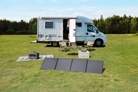 A motorhome is parked on a lush green field surrounded by trees. Nearby, a portable solar panel is set up on the grass beside camping chairs and a table. There is a small power generator and an outdoor rug, creating a comfortable outdoor scene.