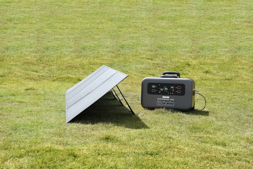 A portable solar panel is angled on a grassy lawn next to a compact power station. The power station displays a digital interface with various indicators.
