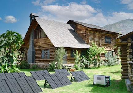 A rustic log cabin with a steeply pitched roof is surrounded by lush green grass and various plants. The cabin has small, barred windows and a chimney. In the foreground, several solar panels are set up on the grass, connected to a portable power generator.