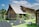A rustic log cabin with a steeply pitched roof is surrounded by lush green grass and various plants. The cabin has small, barred windows and a chimney. In the foreground, several solar panels are set up on the grass, connected to a portable power generator.