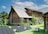 A rustic log cabin with a steeply pitched roof is surrounded by lush green grass and various plants. The cabin has small, barred windows and a chimney. In the foreground, several solar panels are set up on the grass, connected to a portable power generator.