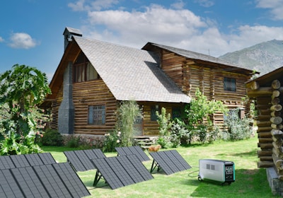 A rustic log cabin with a steeply pitched roof is surrounded by lush green grass and various plants. The cabin has small, barred windows and a chimney. In the foreground, several solar panels are set up on the grass, connected to a portable power generator.