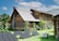 A rustic log cabin with a steeply pitched roof is surrounded by lush green grass and various plants. The cabin has small, barred windows and a chimney. In the foreground, several solar panels are set up on the grass, connected to a portable power generator.
