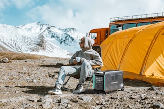 A person in a grey hoodie and pants sits on a folding chair holding a cup. Beside them is a portable power station. Behind them is a large orange tent and an orange vehicle. The background features snow-covered mountains under a partly cloudy sky.