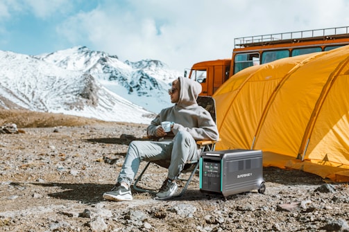 A person in a grey hoodie and pants sits on a folding chair holding a cup. Beside them is a portable power station. Behind them is a large orange tent and an orange vehicle. The background features snow-covered mountains under a partly cloudy sky.