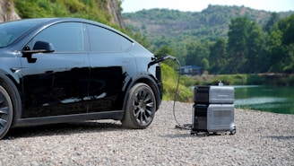 A black electric car is parked on a gravel path by a scenic area with hills and trees in the background. The car is connected to a portable charging unit with a cable.