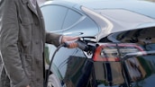 A person wearing a green jacket is holding a charging cable and plugging it into an electric car, specifically at the rear section near the taillight.