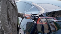 A person wearing a green jacket is holding a charging cable and plugging it into an electric car, specifically at the rear section near the taillight.