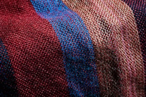 A close-up view of a textured fabric featuring a woven pattern. The fabric showcases alternating stripes in shades of red, blue, and beige, highlighting the fine interlacing of threads.
