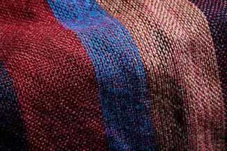 A close-up view of a textured fabric featuring a woven pattern. The fabric showcases alternating stripes in shades of red, blue, and beige, highlighting the fine interlacing of threads.