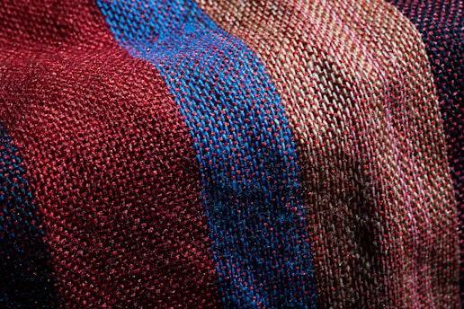 A close-up view of a textured fabric featuring a woven pattern. The fabric showcases alternating stripes in shades of red, blue, and beige, highlighting the fine interlacing of threads.