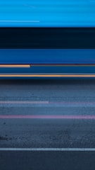 A blurred motion effect creates a dynamic scene with streaks of light and color across a road surface. The image gives a sense of speed, as lines and colors blend together horizontally, reflecting motion and possibly passing vehicles.