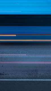 A blurred motion effect creates a dynamic scene with streaks of light and color across a road surface. The image gives a sense of speed, as lines and colors blend together horizontally, reflecting motion and possibly passing vehicles.