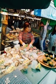 A woman working at a seafood market stall displaying a variety of fresh oysters and other seafood items on ice. The stall is well-lit with overhead lights, and the seafood products are labeled with price tags. There are two other individuals observing or interacting with the stall.