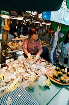A woman working at a seafood market stall displaying a variety of fresh oysters and other seafood items on ice. The stall is well-lit with overhead lights, and the seafood products are labeled with price tags. There are two other individuals observing or interacting with the stall.