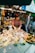 A woman working at a seafood market stall displaying a variety of fresh oysters and other seafood items on ice. The stall is well-lit with overhead lights, and the seafood products are labeled with price tags. There are two other individuals observing or interacting with the stall.