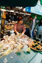 A woman working at a seafood market stall displaying a variety of fresh oysters and other seafood items on ice. The stall is well-lit with overhead lights, and the seafood products are labeled with price tags. There are two other individuals observing or interacting with the stall.