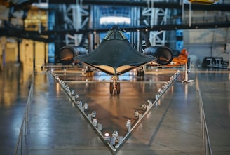 A sleek, black aircraft is prominently displayed in a museum setting. Its design is aerodynamic and futuristic, positioned on a triangular base with lights lining the path leading to it. The surrounding area features industrial-style architecture with metallic textures and structural beams.