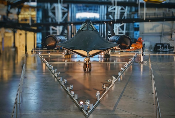 A sleek, black aircraft is prominently displayed in a museum setting. Its design is aerodynamic and futuristic, positioned on a triangular base with lights lining the path leading to it. The surrounding area features industrial-style architecture with metallic textures and structural beams.
