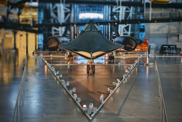 A sleek, black aircraft is prominently displayed in a museum setting. Its design is aerodynamic and futuristic, positioned on a triangular base with lights lining the path leading to it. The surrounding area features industrial-style architecture with metallic textures and structural beams.