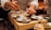 A chef is carefully plating a dish with precision, using chopsticks to add garnish on small plates with meticulously arranged food. The setting is a warm, dimly lit kitchen, suggesting an intimate dining experience.