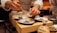 A chef is carefully plating a dish with precision, using chopsticks to add garnish on small plates with meticulously arranged food. The setting is a warm, dimly lit kitchen, suggesting an intimate dining experience.