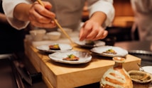 A chef is carefully plating a dish with precision, using chopsticks to add garnish on small plates with meticulously arranged food. The setting is a warm, dimly lit kitchen, suggesting an intimate dining experience.