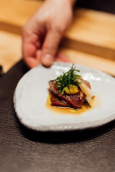 A hand holding a white ceramic plate with a dish composed of sliced meat, garnished with greens and vegetables, and drizzled with a sauce.