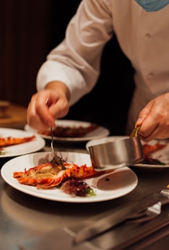 A chef is carefully plating a dish with lobster tails on a white plate, adding a sauce using a metal pot. The focus is on meticulous food presentation in a professional kitchen setting.