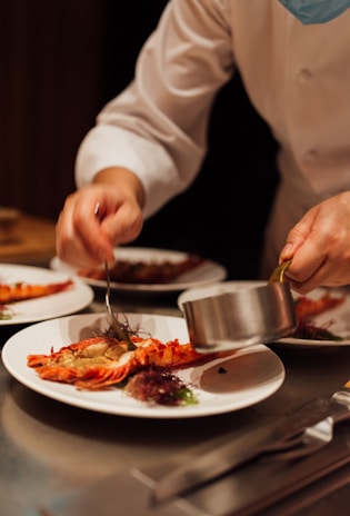 A chef is carefully plating a dish with lobster tails on a white plate, adding a sauce using a metal pot. The focus is on meticulous food presentation in a professional kitchen setting.