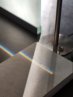 A light grey book, labeled 'Bullet Journal,' is placed on a dark surface. A thin, vibrant rainbow beam of light cuts diagonally across the book cover. The book is next to a metallic lamp with a cylindrical base, and light from an adjacent translucent glass panel creates soft reflections.