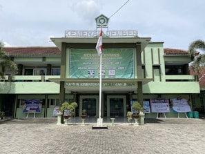 A government building with a large green banner welcoming visitors. The facade features a combination of light green and earthy tones, and there are potted plants flanking the entrance. An Indonesian flag is displayed prominently at the front, and the text above identifies it as the Ministry of Religious Affairs in Jember.