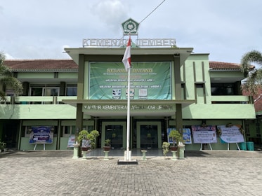 A government building with a large green banner welcoming visitors. The facade features a combination of light green and earthy tones, and there are potted plants flanking the entrance. An Indonesian flag is displayed prominently at the front, and the text above identifies it as the Ministry of Religious Affairs in Jember.