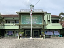 A government building with a large green banner welcoming visitors. The facade features a combination of light green and earthy tones, and there are potted plants flanking the entrance. An Indonesian flag is displayed prominently at the front, and the text above identifies it as the Ministry of Religious Affairs in Jember.