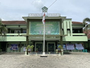 A government building with a large green banner welcoming visitors. The facade features a combination of light green and earthy tones, and there are potted plants flanking the entrance. An Indonesian flag is displayed prominently at the front, and the text above identifies it as the Ministry of Religious Affairs in Jember.