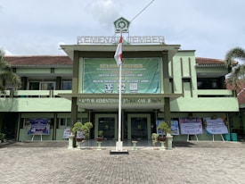 A government building with a large green banner welcoming visitors. The facade features a combination of light green and earthy tones, and there are potted plants flanking the entrance. An Indonesian flag is displayed prominently at the front, and the text above identifies it as the Ministry of Religious Affairs in Jember.