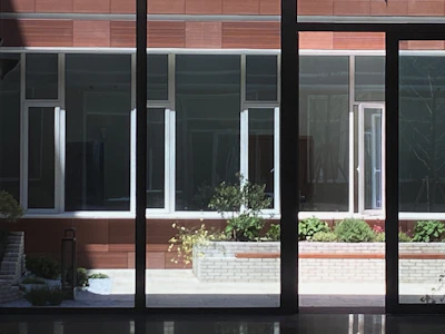 A courtyard view featuring large windows reflecting light, bordered by brown paneling. A central planter contains green shrubs and small plants with a brick border.