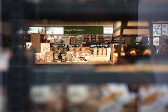 A store shelf filled with various packaged snacks and paper products. The foreground contains bags and cans, while in the background, shelves hold a variety of items including boxed cereals and napkins. Shoppers can be seen in the aisle beyond the shelves.
