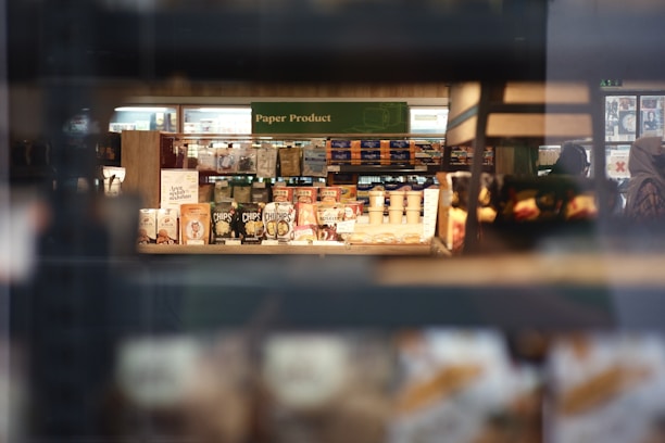 A store shelf filled with various packaged snacks and paper products. The foreground contains bags and cans, while in the background, shelves hold a variety of items including boxed cereals and napkins. Shoppers can be seen in the aisle beyond the shelves.