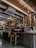 A cozy coffee shop interior with a rustic design, featuring a wooden ceiling and walls, pendant lights, and a modern espresso machine on a black counter. Two stylish wooden bar stools are placed in front of the counter. The shelves in the background are lined with several coffee and kitchen items, contributing to the warm and inviting atmosphere.