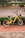 A yellow rental bicycle with a black basket is parked on a red pedestrian pathway near a street with a low hedge in the background. The bicycle is positioned against a backdrop of green foliage and a paved road, while a partially blurry car is visible driving by.