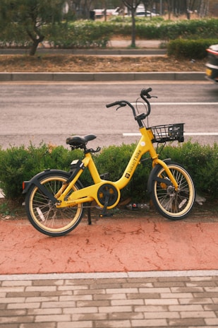 A yellow rental bicycle with a black basket is parked on a red pedestrian pathway near a street with a low hedge in the background. The bicycle is positioned against a backdrop of green foliage and a paved road, while a partially blurry car is visible driving by.