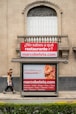 A street advertisement is placed in front of a stone building with an arched window. The ad promotes a website, marcobeteta.com, and suggests finding restaurants. It lists various cuisines such as Mexican, Israeli, Greek, and more, with an image of food including pasta and what appears to be shrimp.