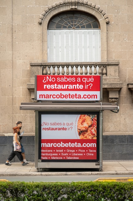 A street advertisement is placed in front of a stone building with an arched window. The ad promotes a website, marcobeteta.com, and suggests finding restaurants. It lists various cuisines such as Mexican, Israeli, Greek, and more, with an image of food including pasta and what appears to be shrimp.