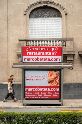 A street advertisement is placed in front of a stone building with an arched window. The ad promotes a website, marcobeteta.com, and suggests finding restaurants. It lists various cuisines such as Mexican, Israeli, Greek, and more, with an image of food including pasta and what appears to be shrimp.